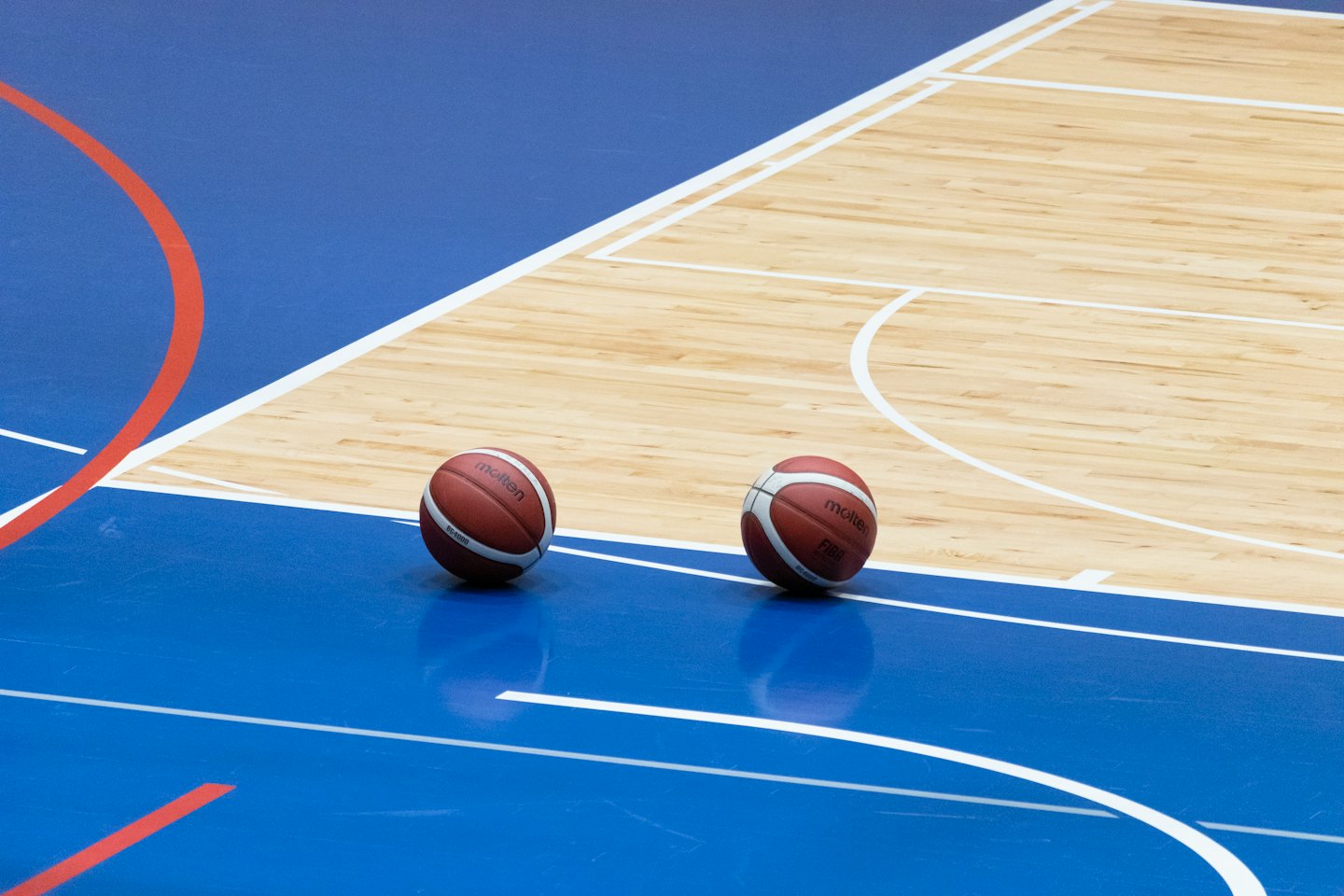 Two basketballs sit on a blue and wood indoor court.